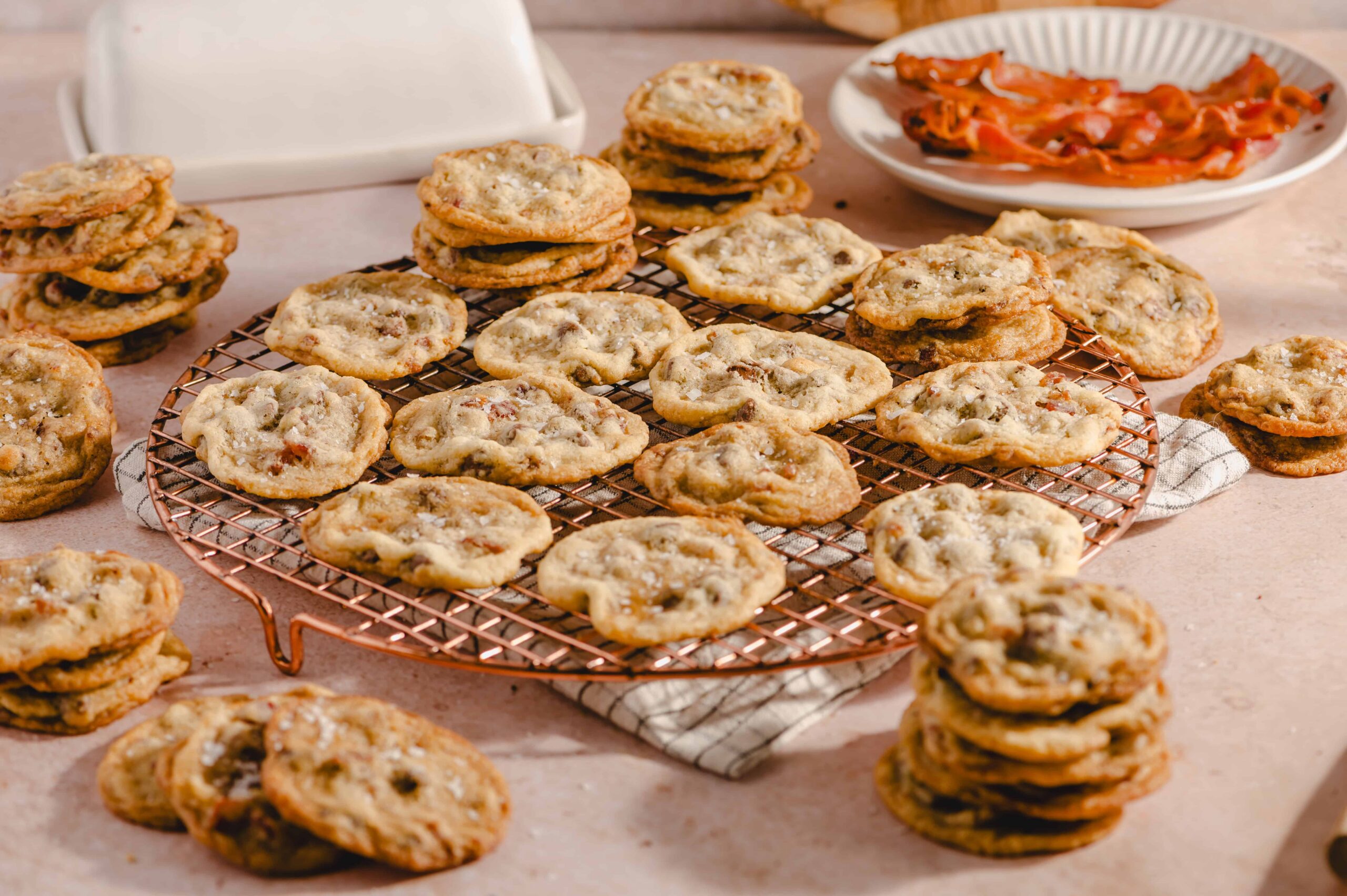Buttery chocolate chip cookies cooling on a wire rack with a plate of crispy bacon in the background.