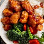 Crispy orange chicken with broccoli and rice in a decorative bowl, garnished with chopped vegetables on a gray textured surface, served with chopsticks for an Asian-inspired meal.