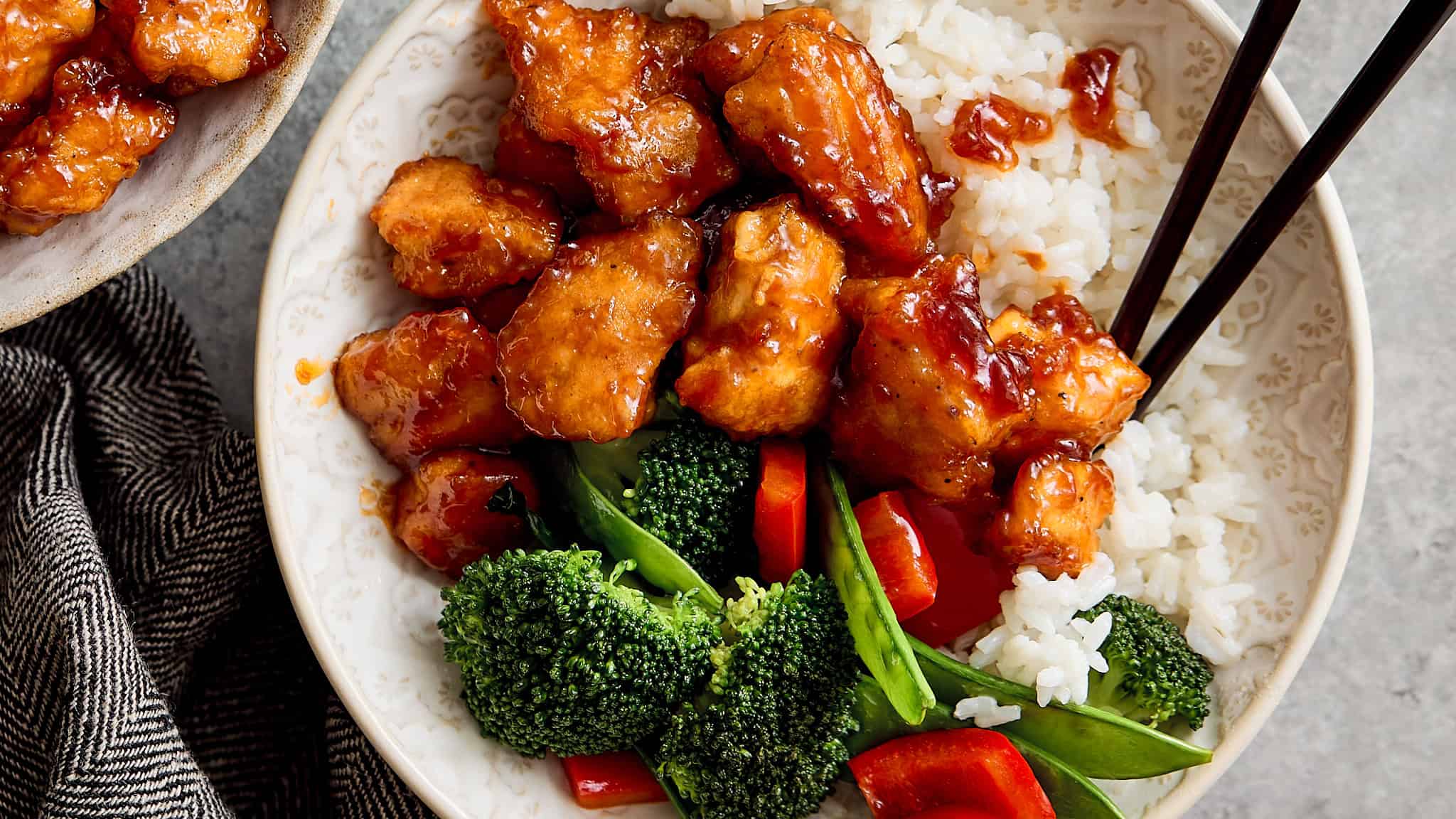 Crispy orange chicken with broccoli and rice in a decorative bowl, garnished with chopped vegetables on a gray textured surface, served with chopsticks for an Asian-inspired meal.
