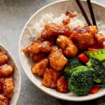 Crispy orange chicken with vegetables served over steamed white rice in a decorative bowl, garnished with broccoli, red bell peppers, and snap peas, perfect for Asian-inspired dinner or takeout.