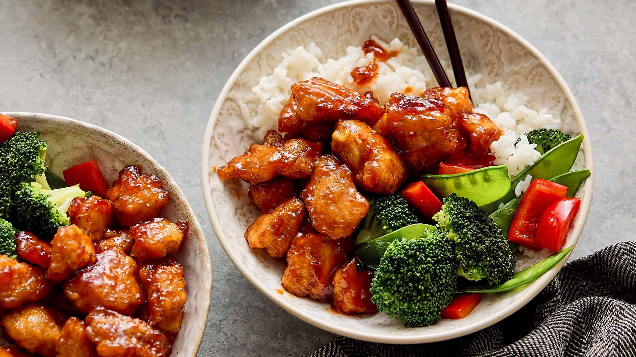 Crispy orange chicken with vegetables served over steamed white rice in a decorative bowl, garnished with broccoli, red bell peppers, and snap peas, perfect for Asian-inspired dinner or takeout.