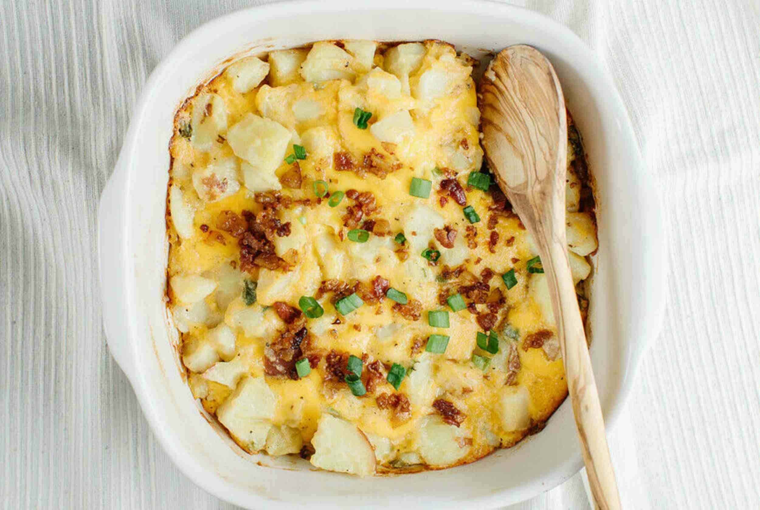 Baked potato casserole with cheese, bacon, and green onions in a white dish, wooden spoon for serving, comforting potato dish, easy casserole recipe, hearty comfort food.