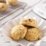 Soft savory scones with cheese and herbs in a white fluted bowl, gluten-friendly baked breakfast treat, ideal for brunch or snacks, served on a white table with baking tray and ingredients in the background.