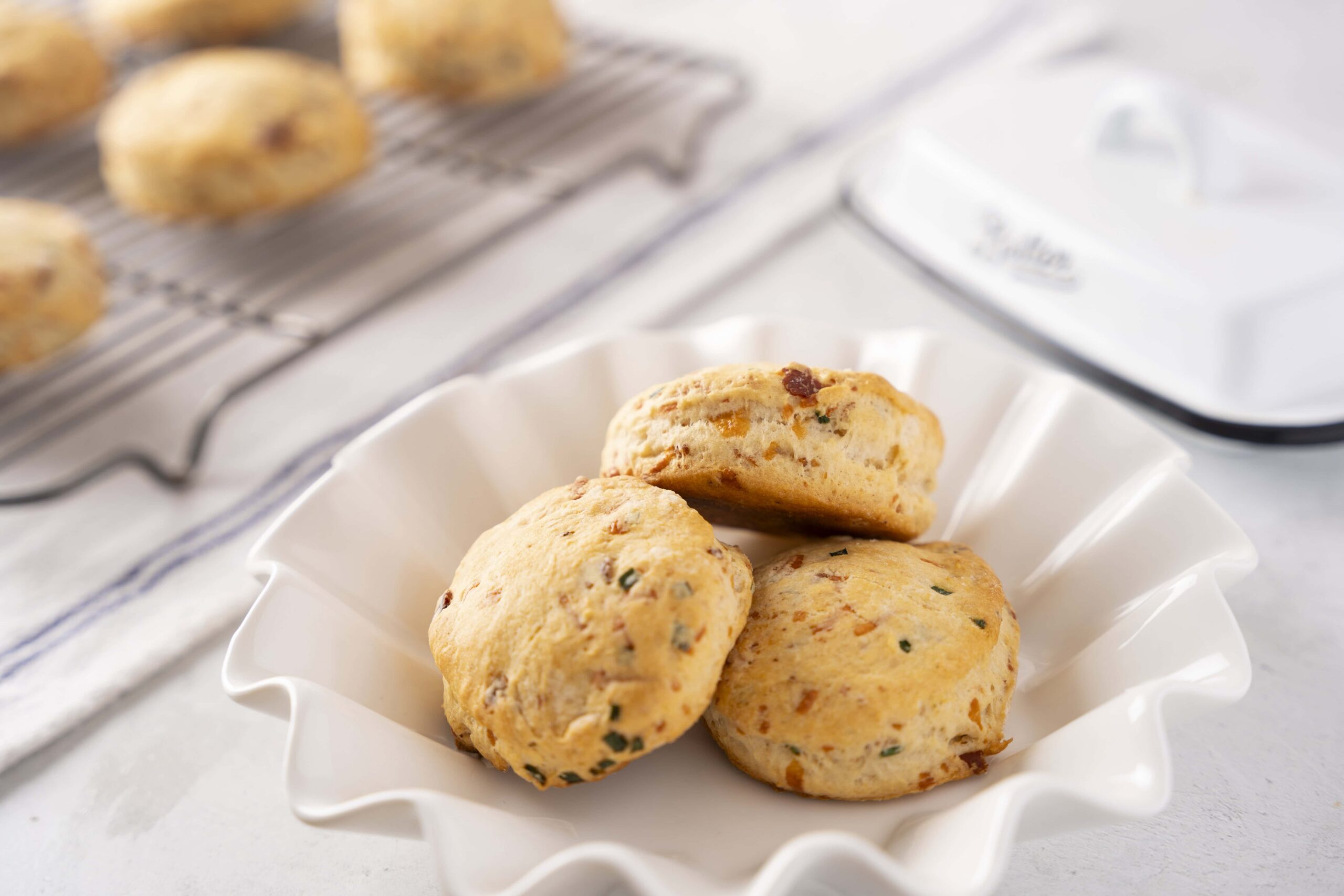 Soft savory scones with cheese and herbs in a white fluted bowl, gluten-friendly baked breakfast treat, ideal for brunch or snacks, served on a white table with baking tray and ingredients in the background.