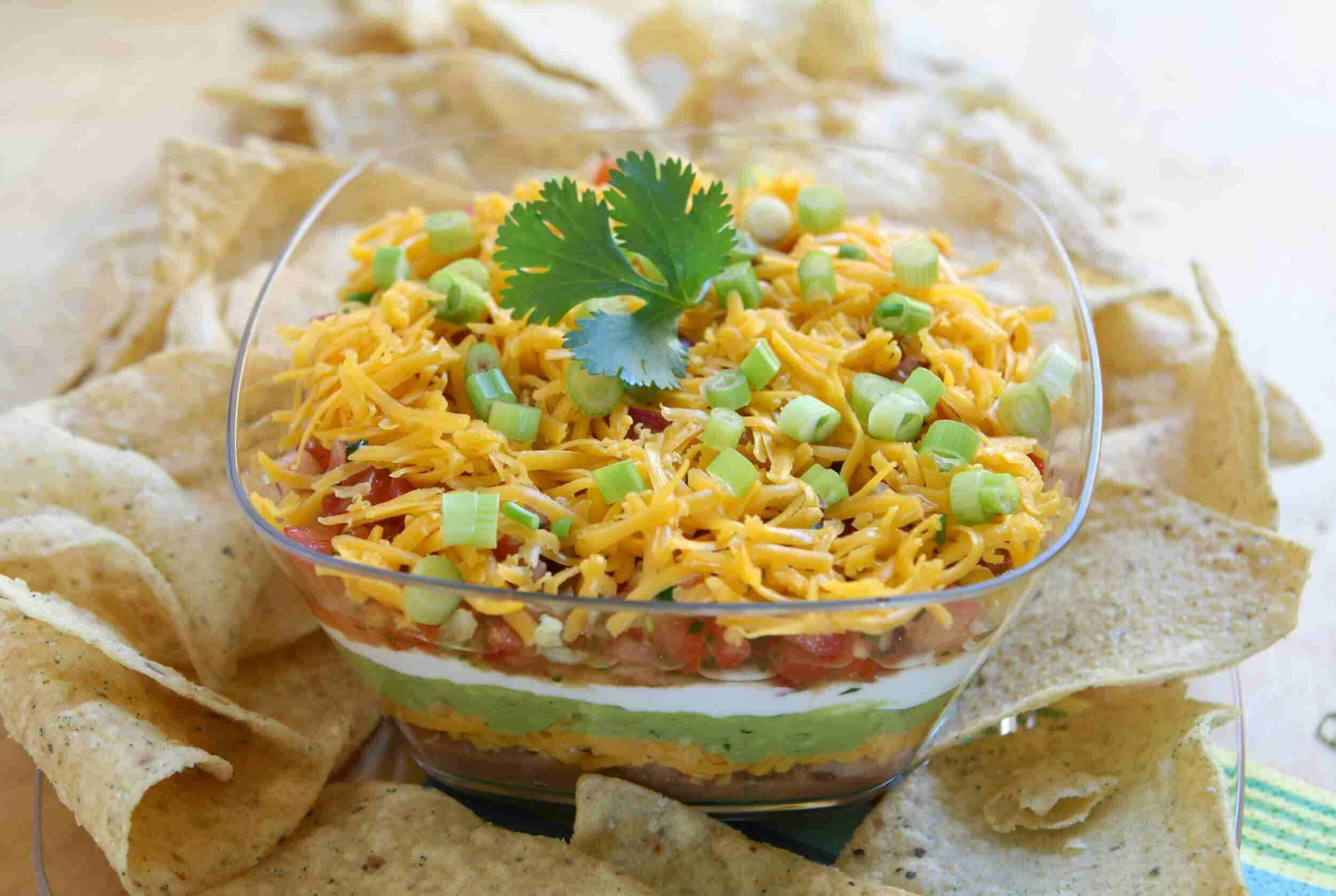 Fresh layered taco dip with shredded cheese, chopped green onions, and cilantro in a glass bowl, surrounded by tortilla chip scoops, perfect for parties and snack platters.
