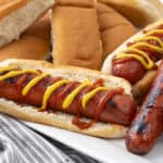 Hot dogs with mustard and ketchup in a soft bun, served alongside grilled sausages and sliced bread on a white plate.