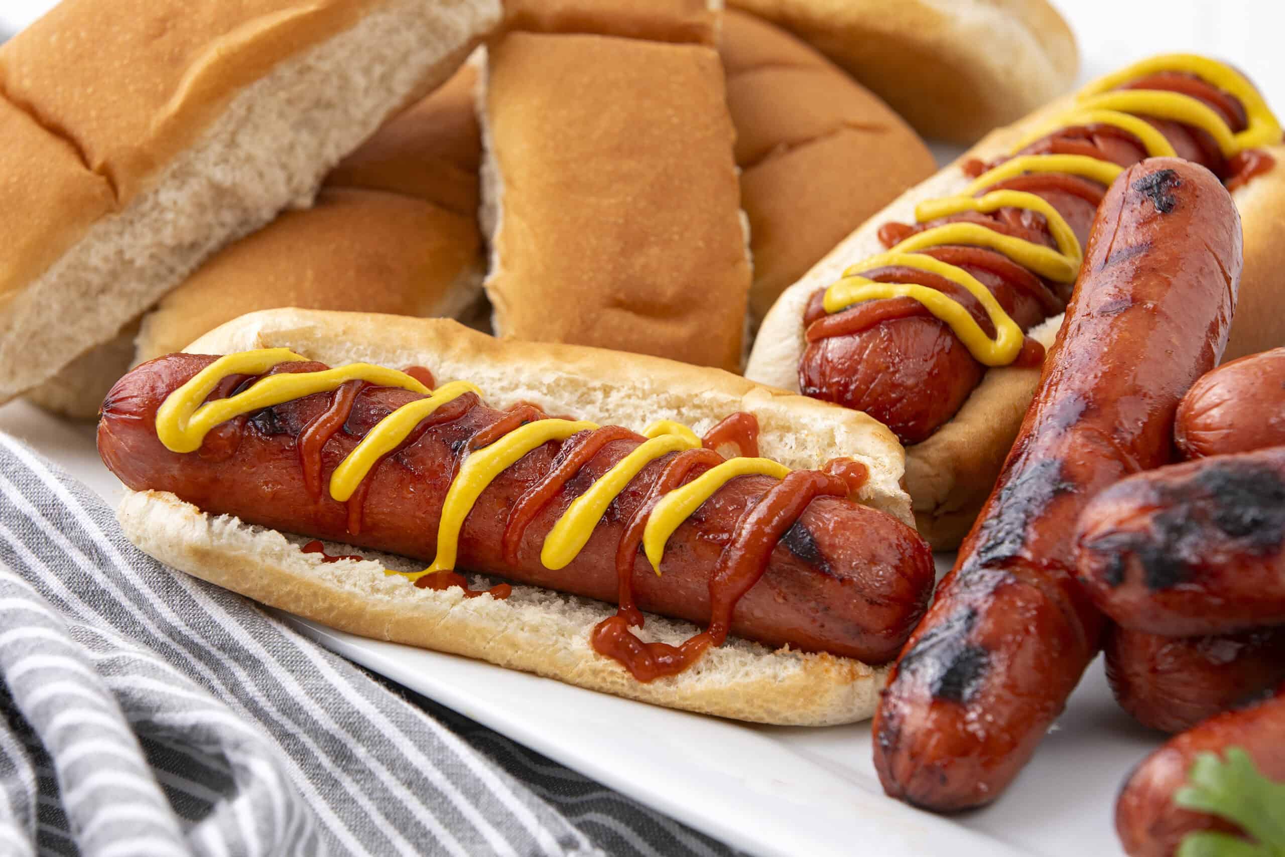Hot dogs with mustard and ketchup in a soft bun, served alongside grilled sausages and sliced bread on a white plate.