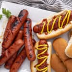 Grilled sausages and hot dogs with mustard and ketchup served on a white platter, featuring soft buns and hot dog rolls, perfect for a classic barbecue or hot dog meal.