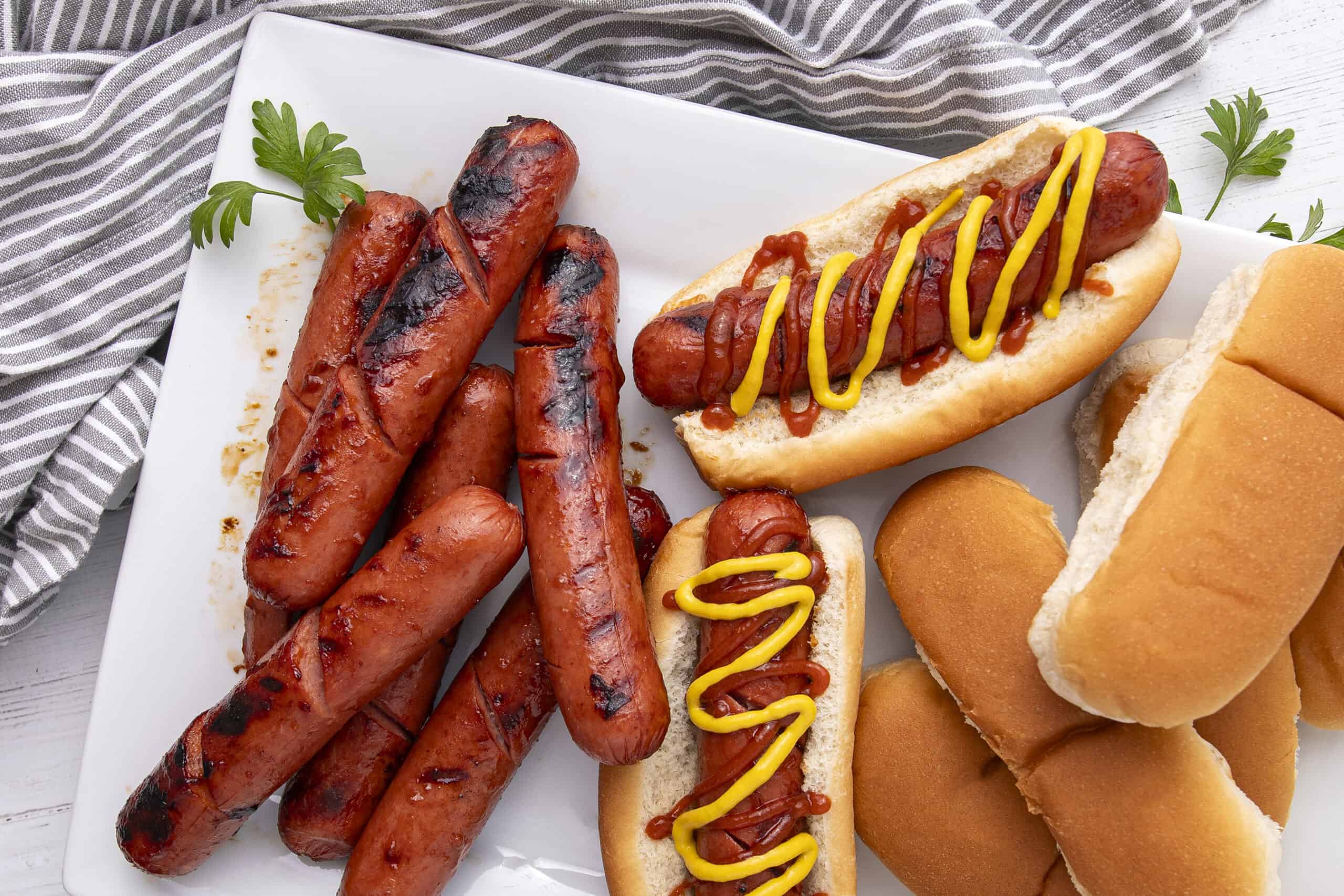 Grilled sausages and hot dogs with mustard and ketchup served on a white platter, featuring soft buns and hot dog rolls, perfect for a classic barbecue or hot dog meal.