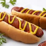 Hot dog with grilled sausages, topped with mustard and ketchup in a soft bun, served on a white plate with fresh parsley garnish.