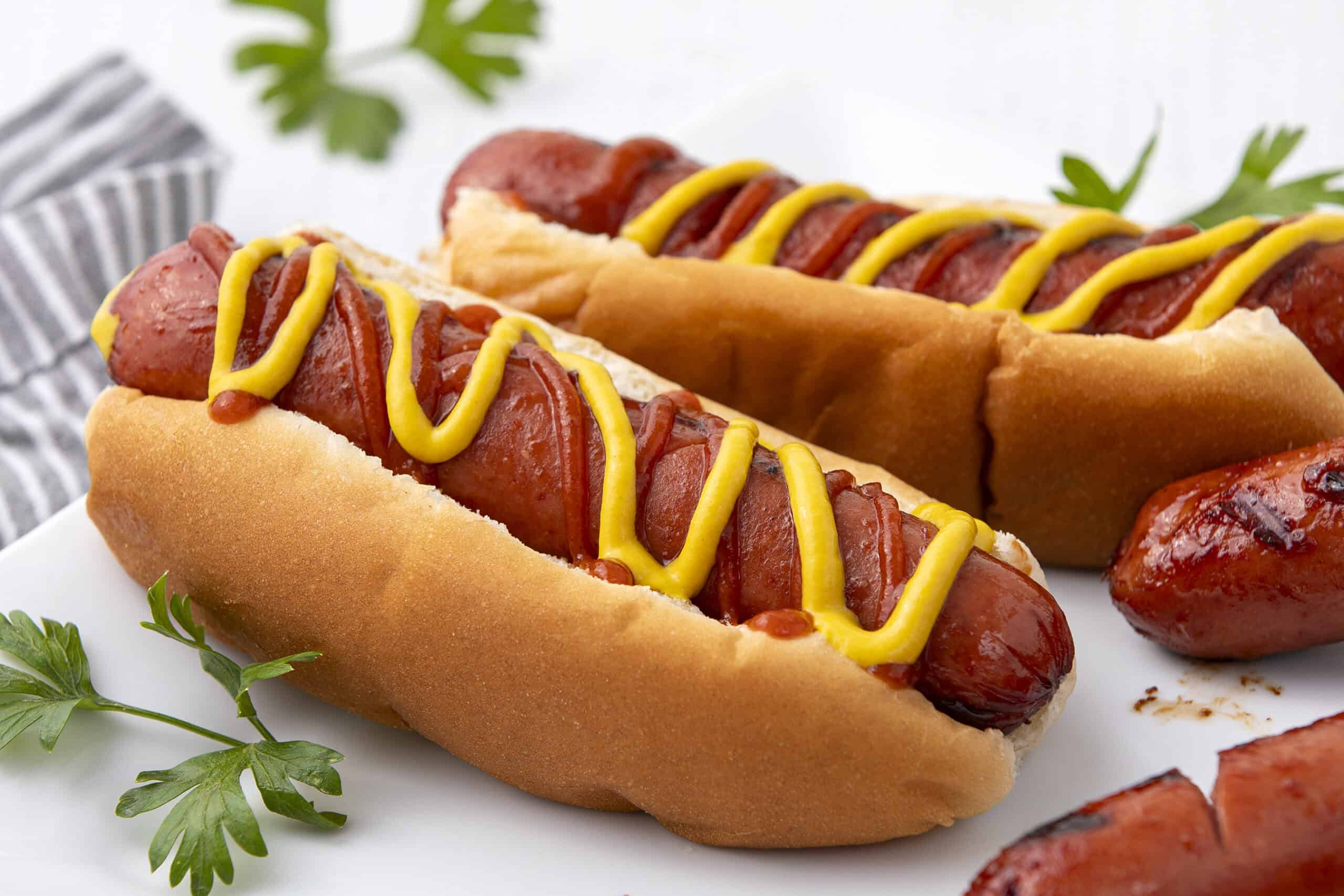 Hot dog with grilled sausages, topped with mustard and ketchup in a soft bun, served on a white plate with fresh parsley garnish.