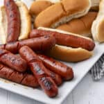 Grilled hot dogs with toasted buns and sausages on a white serving platter, surrounded by fresh bread rolls, ideal for barbecue or quick meals.