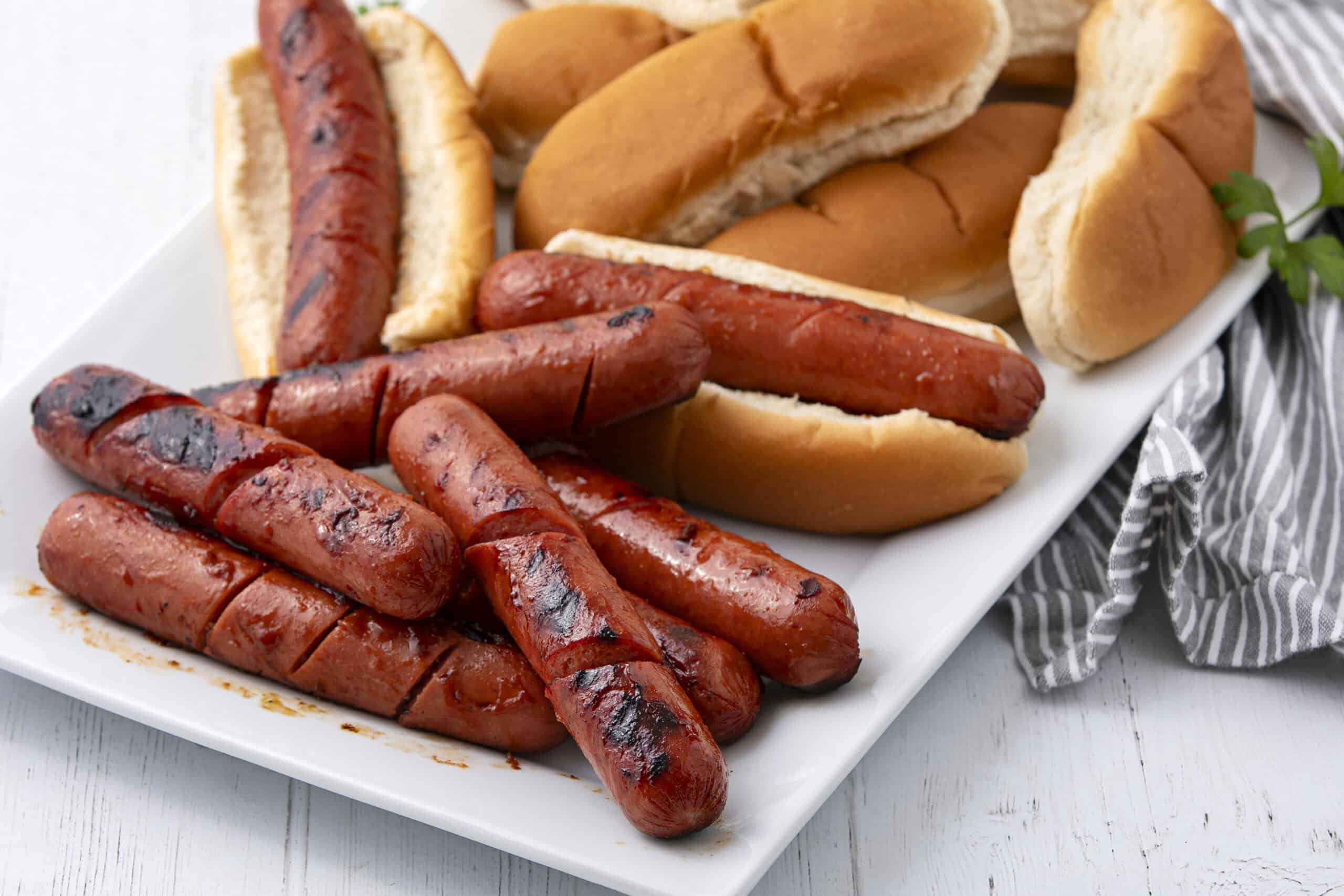 Grilled hot dogs with toasted buns and sausages on a white serving platter, surrounded by fresh bread rolls, ideal for barbecue or quick meals.