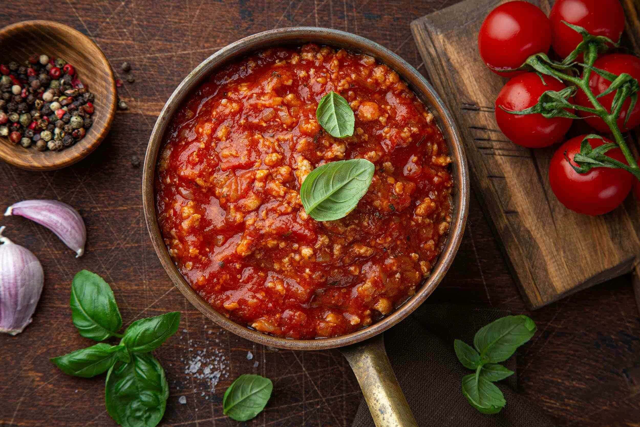 Rich homemade spaghetti bolognese in a wooden bowl with fresh basil on top, surrounded by cherry tomatoes, garlic, and peppercorns on a rustic wooden surface.