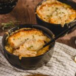Creamy chicken pot pie dessert with melted cheese in black bowls on rustic wooden table with gold spoons and salt shaker.