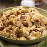 Creamy bacon and egg pasta salad in a light green bowl with crispy bacon bits on top, served on a rustic wooden table with a colorful cloth underneath. Ideal for summer recipes or hearty side dishes.