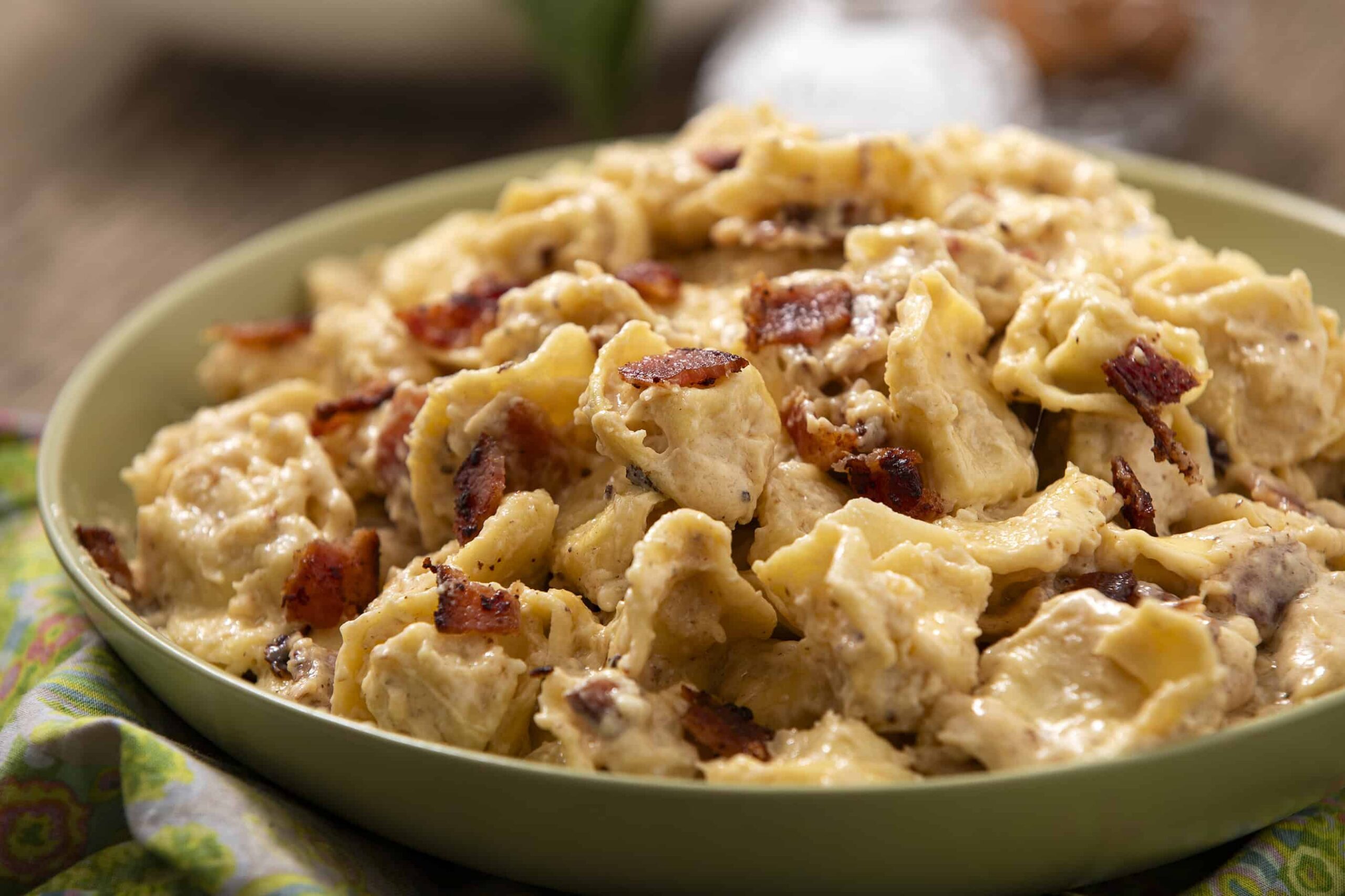 Savory creamy chicken and bacon pasta in a green bowl, topped with crispy bacon bits, served on a colorful cloth. Perfect comfort food for a hearty meal.