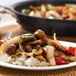 Steamed rice topped with savory stir-fried vegetables and tender pieces of meat, served on a white plate with a flavorful sauce and colorful peppers, showcasing an Asian-inspired dish.