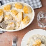 Creamy mashed potatoes with gravy and chunks of chicken in a white casserole dish, served with a glass of water and a small plate with a scoop of mashed potatoes, on a rustic table.
