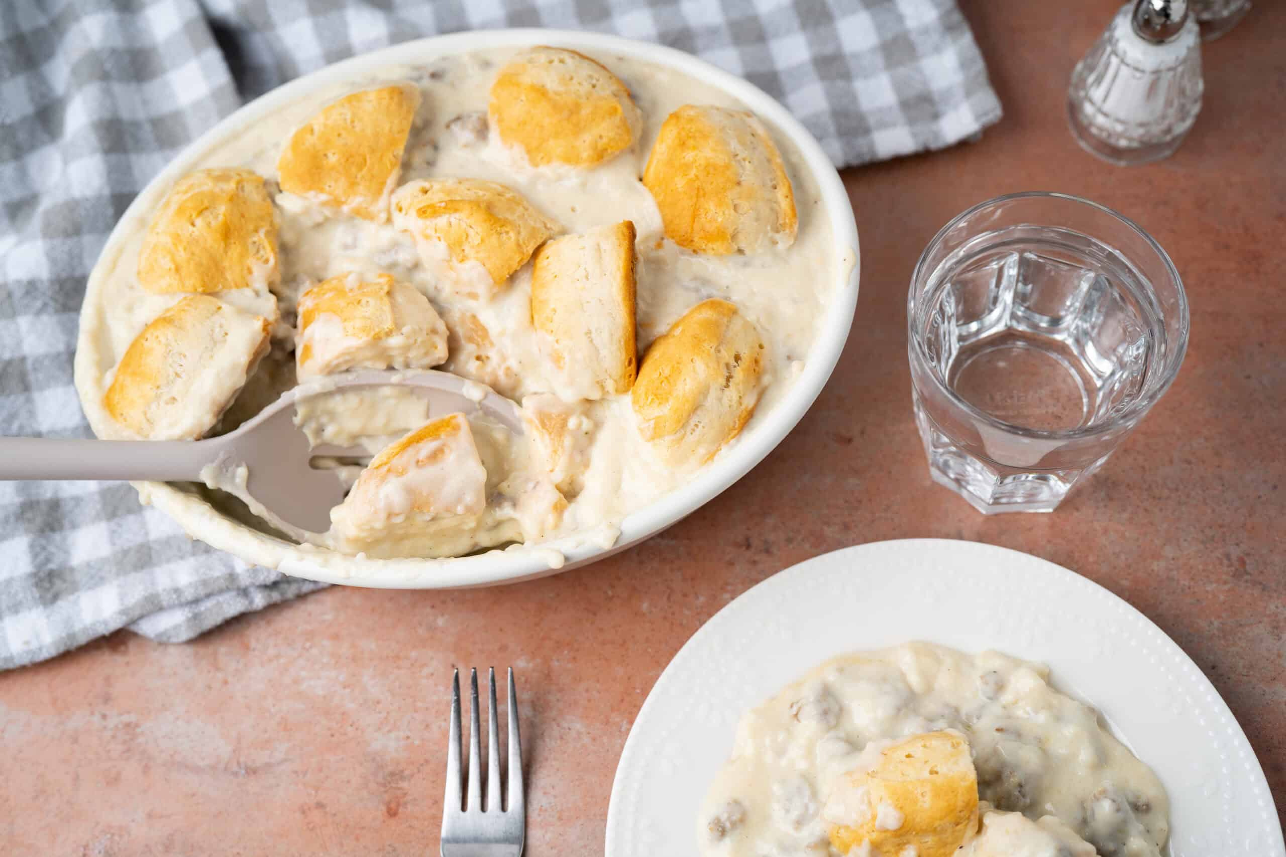 Creamy mashed potatoes with gravy and chunks of chicken in a white casserole dish, served with a glass of water and a small plate with a scoop of mashed potatoes, on a rustic table.