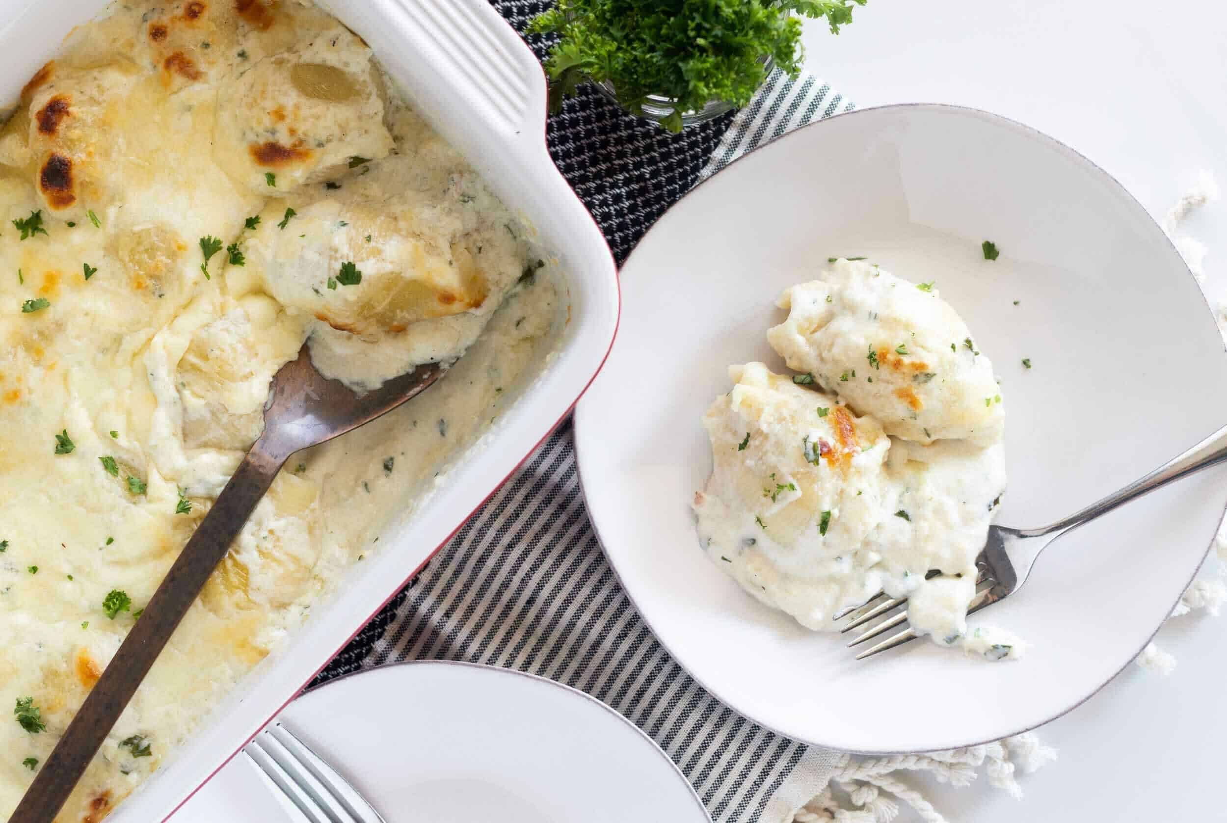 Creamy garlic mashed potatoes with herbs topped with melted cheese, served in a white baking dish and a white plate with a fork, garnished with fresh parsley. Perfect comfort food side dish.