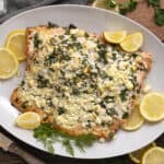 Baked salmon fillet topped with crumbled feta cheese, fresh herbs, and lemon slices on a white platter. Perfect for healthy dinner recipes and seafood lovers.