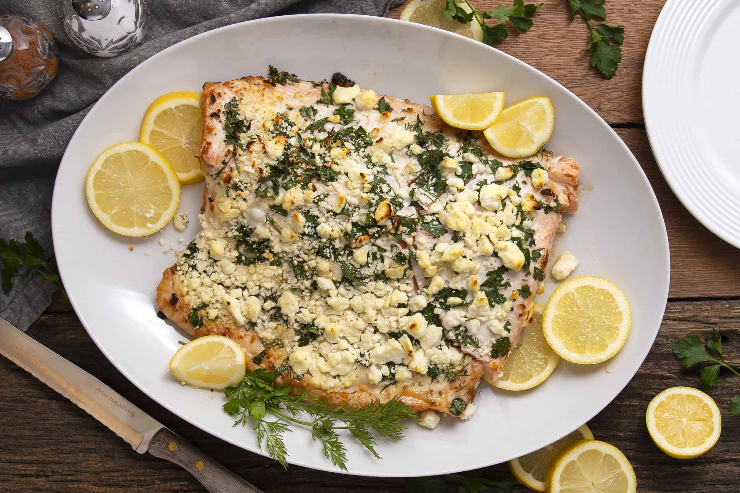 Baked salmon fillet topped with crumbled feta cheese, fresh herbs, and lemon slices on a white platter. Perfect for healthy dinner recipes and seafood lovers.