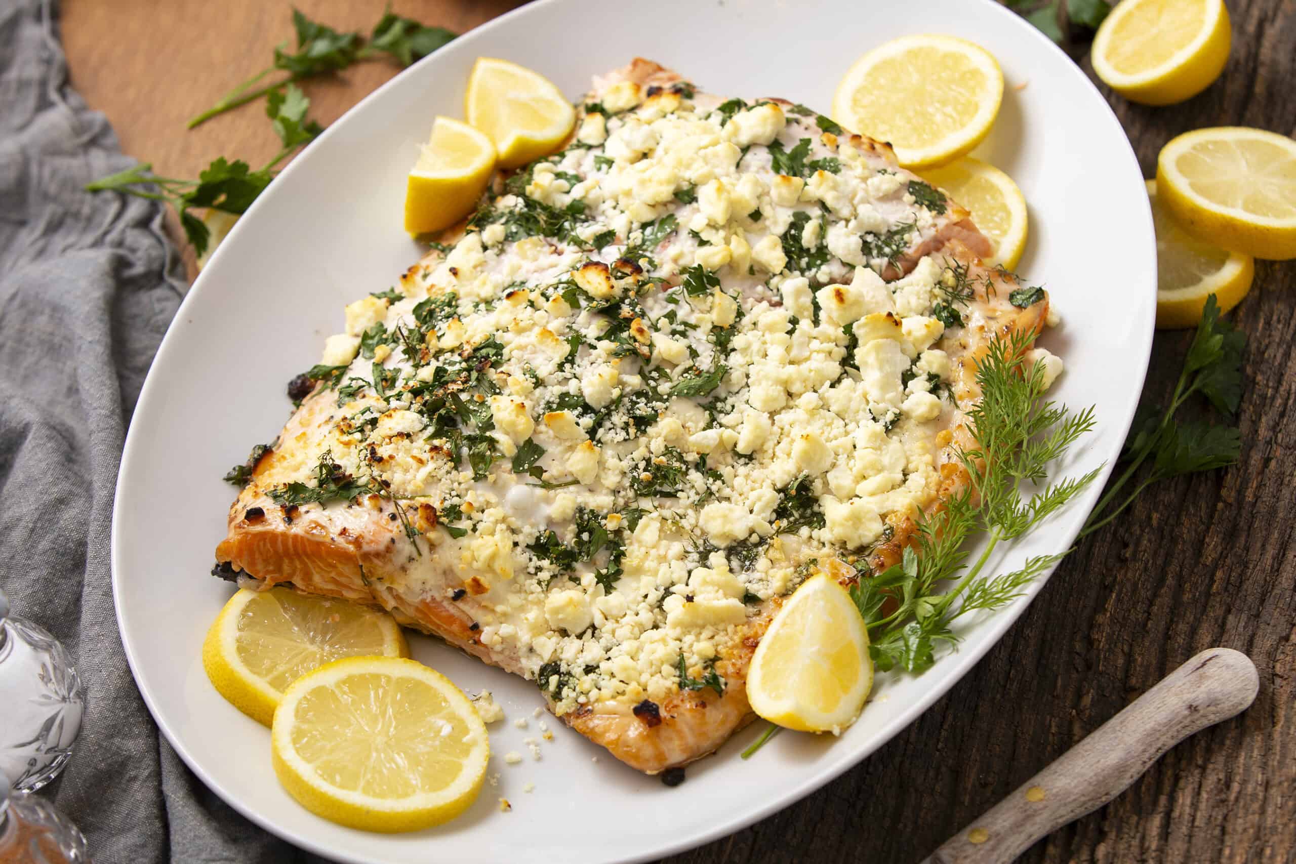 Delicious baked salmon topped with crumbled feta cheese, fresh herbs, and lemon wedges on a white platter. Perfect for a healthy seafood dinner or lunch.