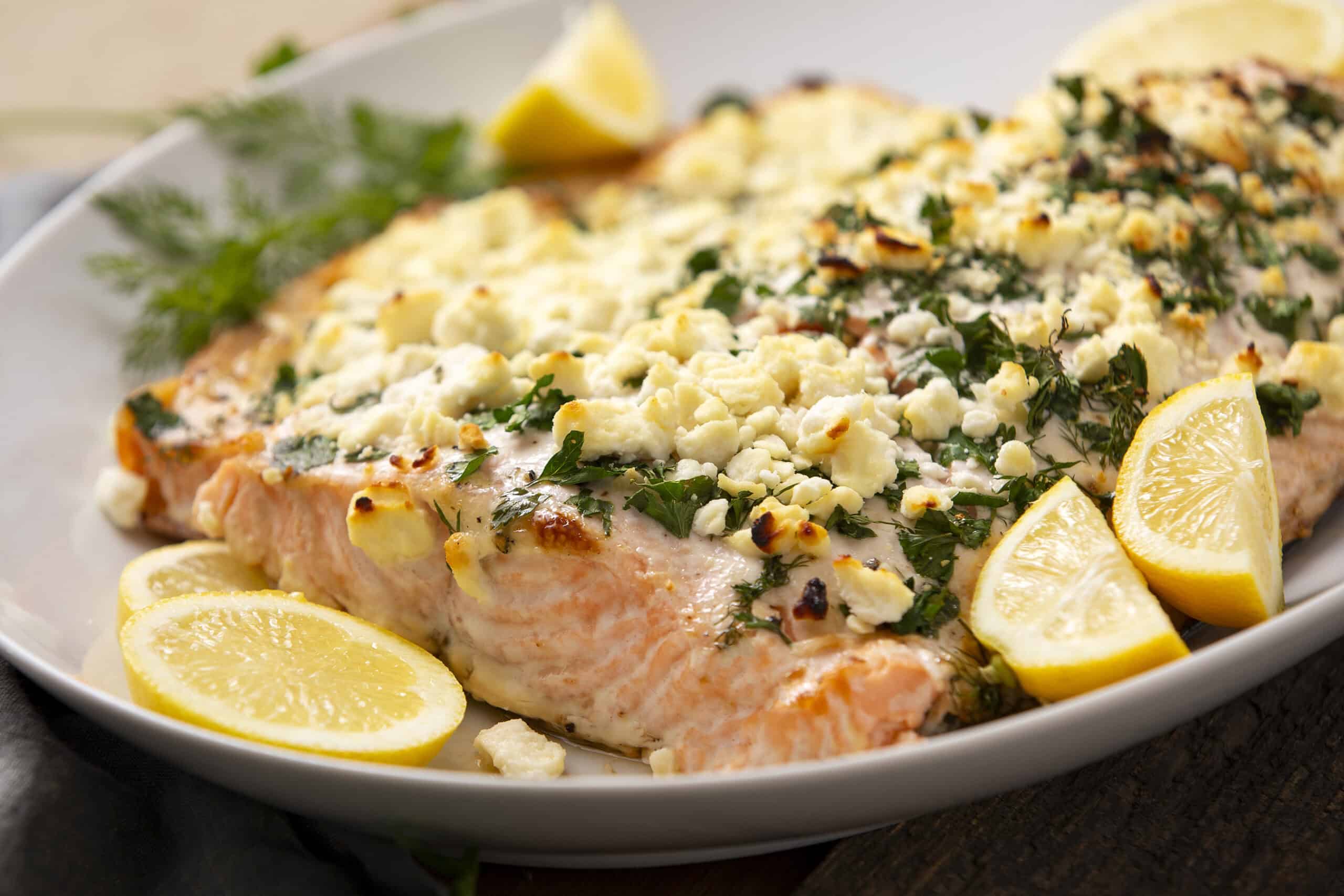 Juicy baked salmon fillet topped with crumbled feta cheese, fresh herbs, and lemon slices on a white platter, perfect for healthy seafood recipes and dinner ideas.