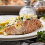 Fresh baked salmon fillet topped with crumbled feta cheese, herbs, and lemon slices on a white plate, served with additional lemon wedges and garnished with parsley, perfect for healthy seafood recipes.