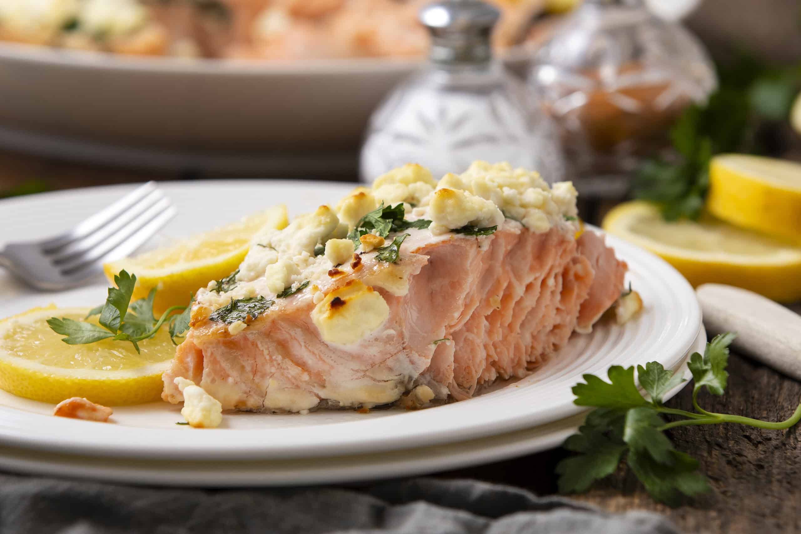 Fresh baked salmon fillet topped with crumbled feta cheese, herbs, and lemon slices on a white plate, served with additional lemon wedges and garnished with parsley, perfect for healthy seafood recipes.