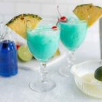 Refreshing blue cocktails garnished with pineapple slices and cherries, served in elegant glasses with ingredients like lime and blue curaçao visible in the background.