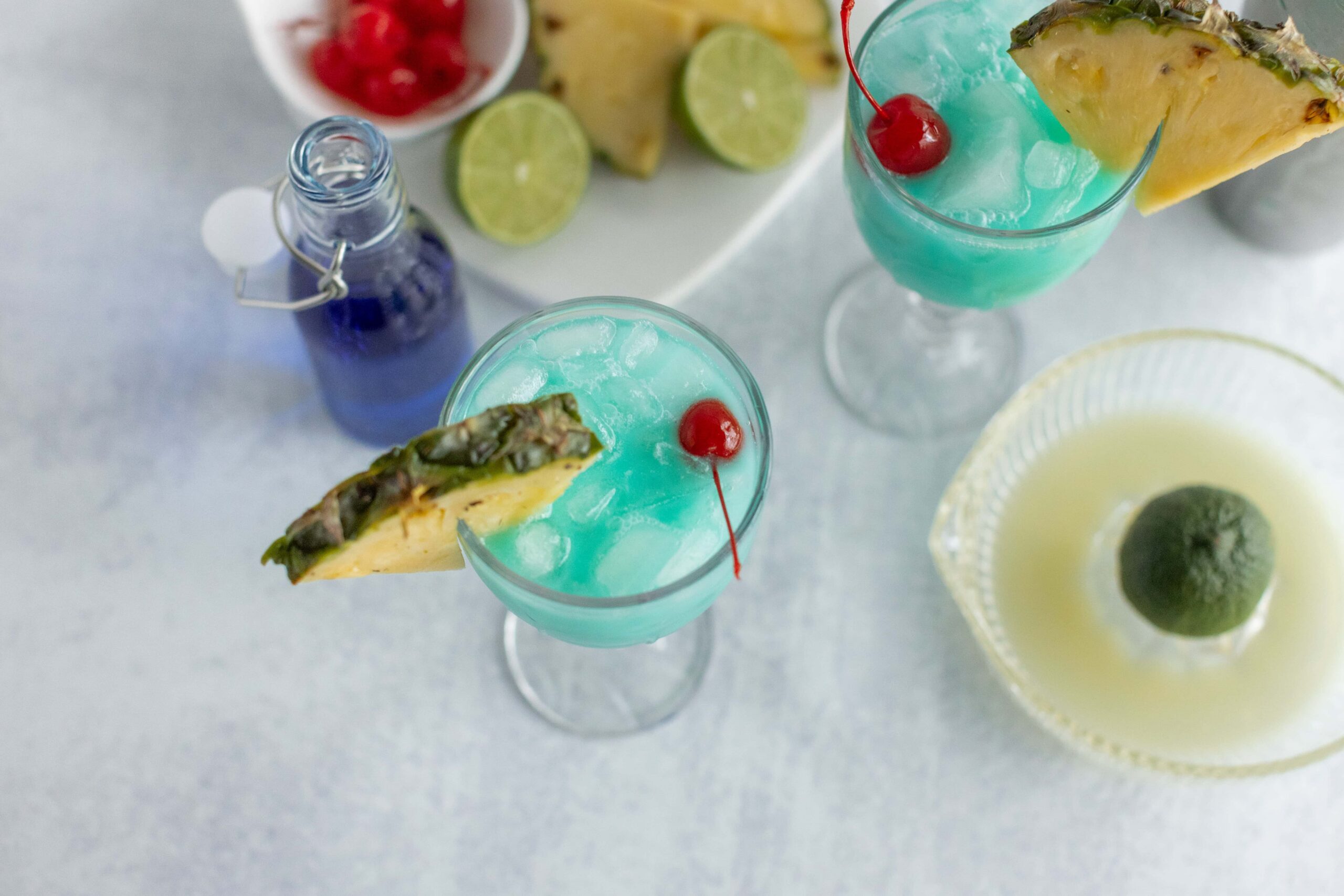 Refreshing blue tropical drinks garnished with pineapple slices and maraschino cherries, served with lime wedges, perfect for summer parties.