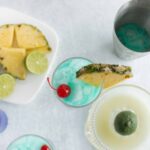 Vibrant blue cocktails garnished with pineapple slices and cherries, served alongside fresh pineapple chunks, lime wedges, and various colorful drinks on a white table for a summery party vibe.