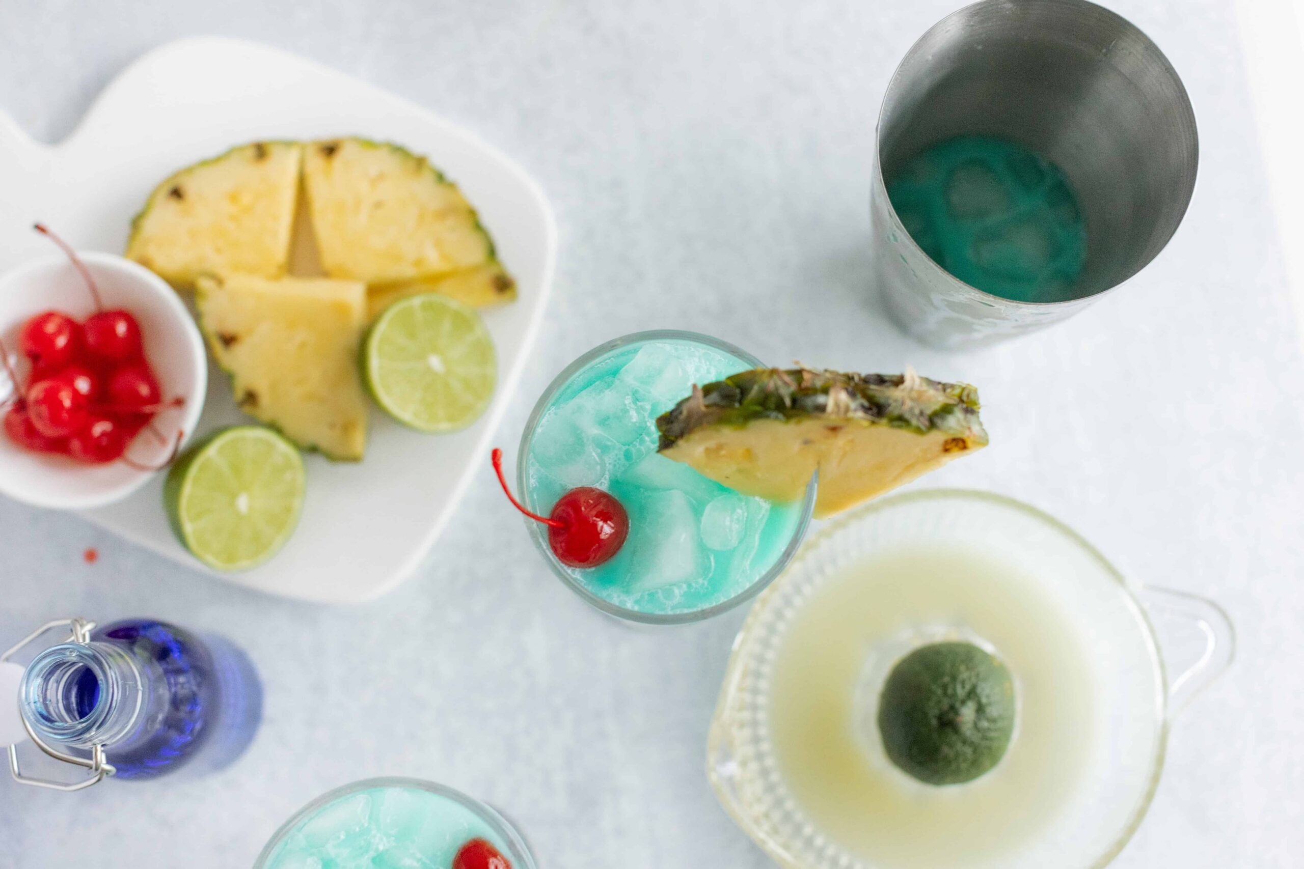 Vibrant blue cocktails garnished with pineapple slices and cherries, served alongside fresh pineapple chunks, lime wedges, and various colorful drinks on a white table for a summery party vibe.