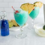 Refreshing blue tropical cocktails garnished with pineapple slices and cherries, served in elegant glasses with ingredients like lime and blue Curaçao on a bright white countertop.