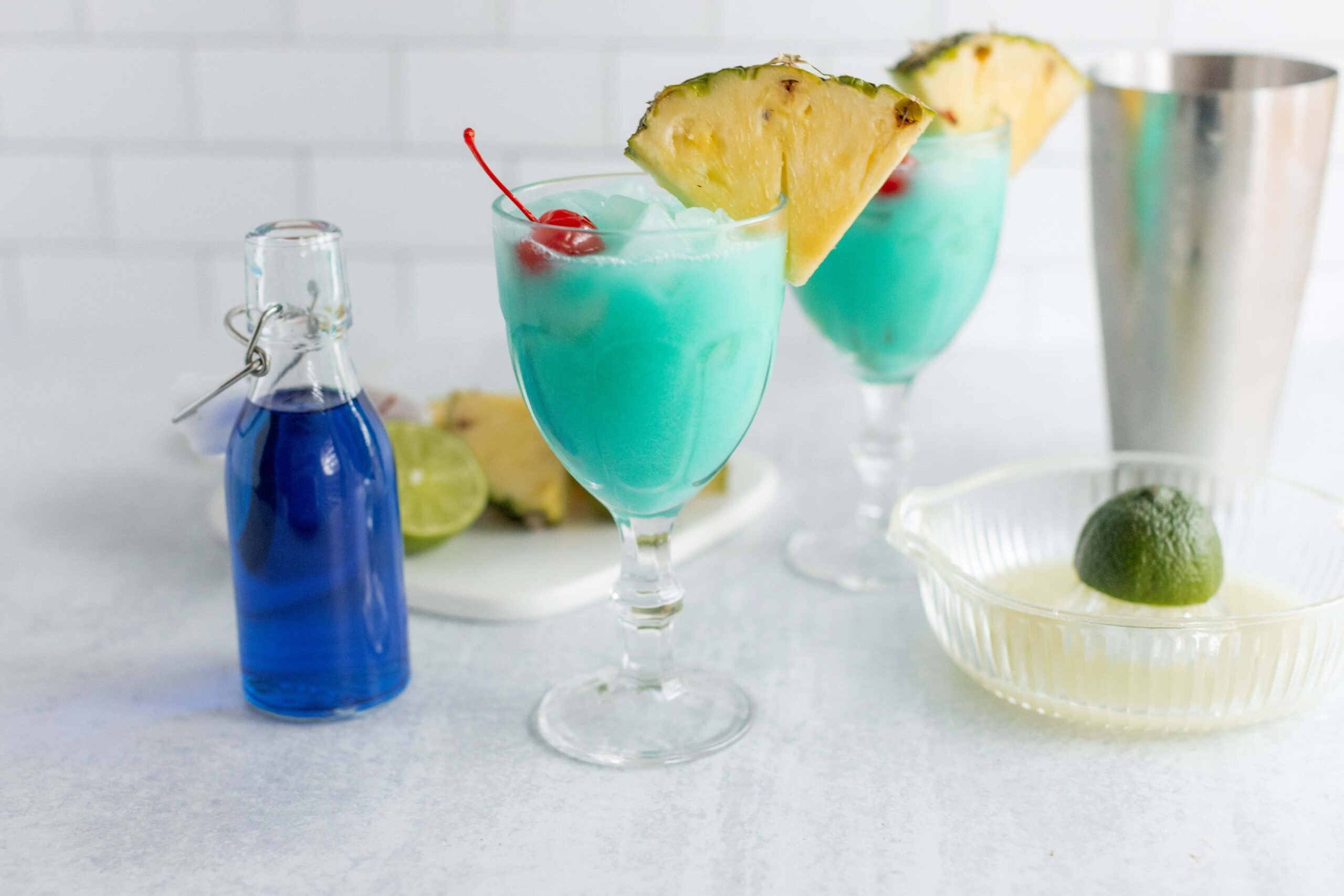 Refreshing blue tropical cocktails garnished with pineapple slices and cherries, served in elegant glasses with ingredients like lime and blue Curaçao on a bright white countertop.