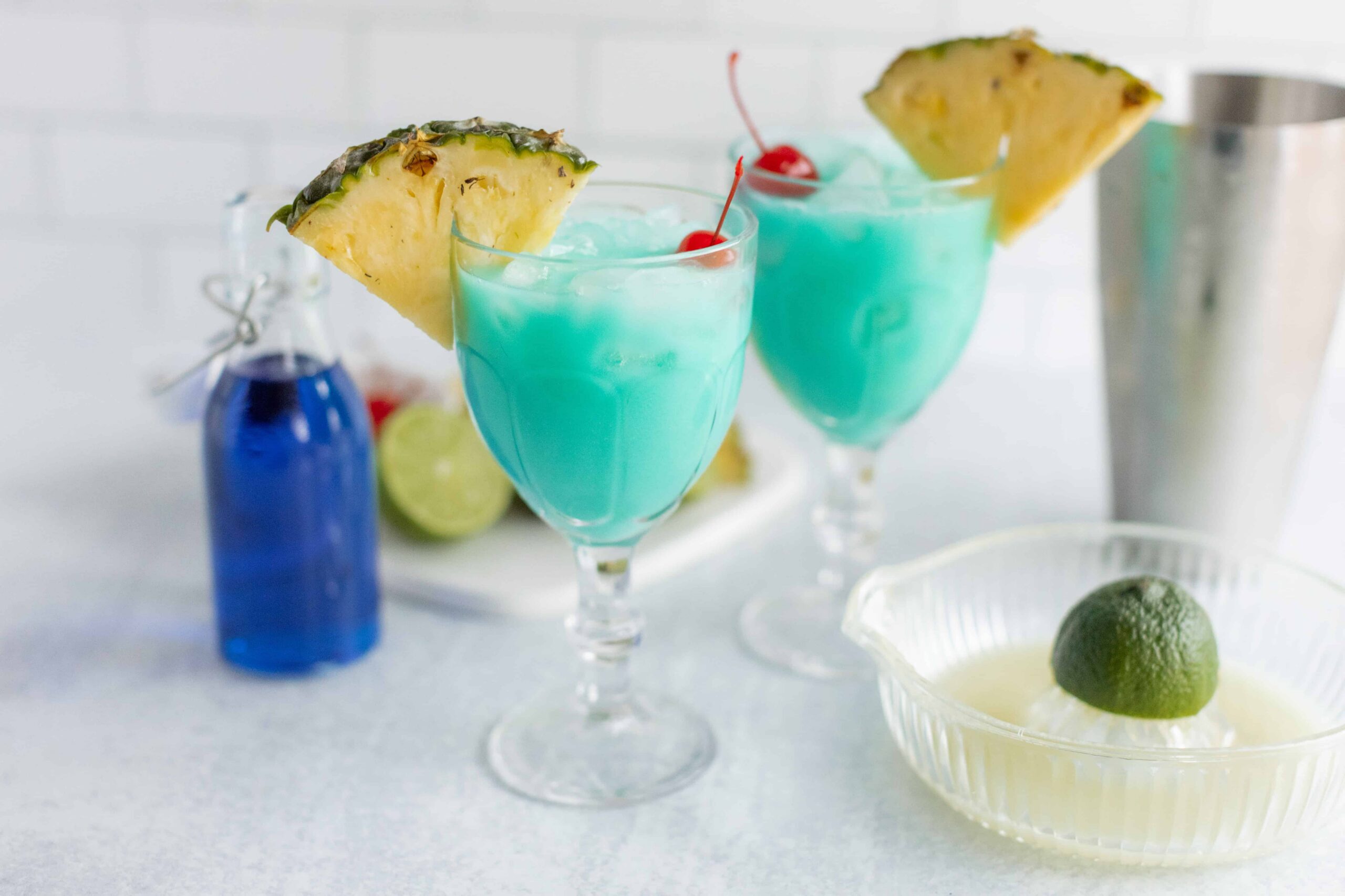 Refreshing blue cocktails garnished with pineapple slices and cherries, served in elegant glasses with ingredients like lime and blue curaçao visible in the background.
