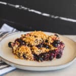 Baked mixed berry crumble on a white plate with a black background and striped cloth, perfect for summer dessert, healthy berry dessert, easy berry crumble recipe.