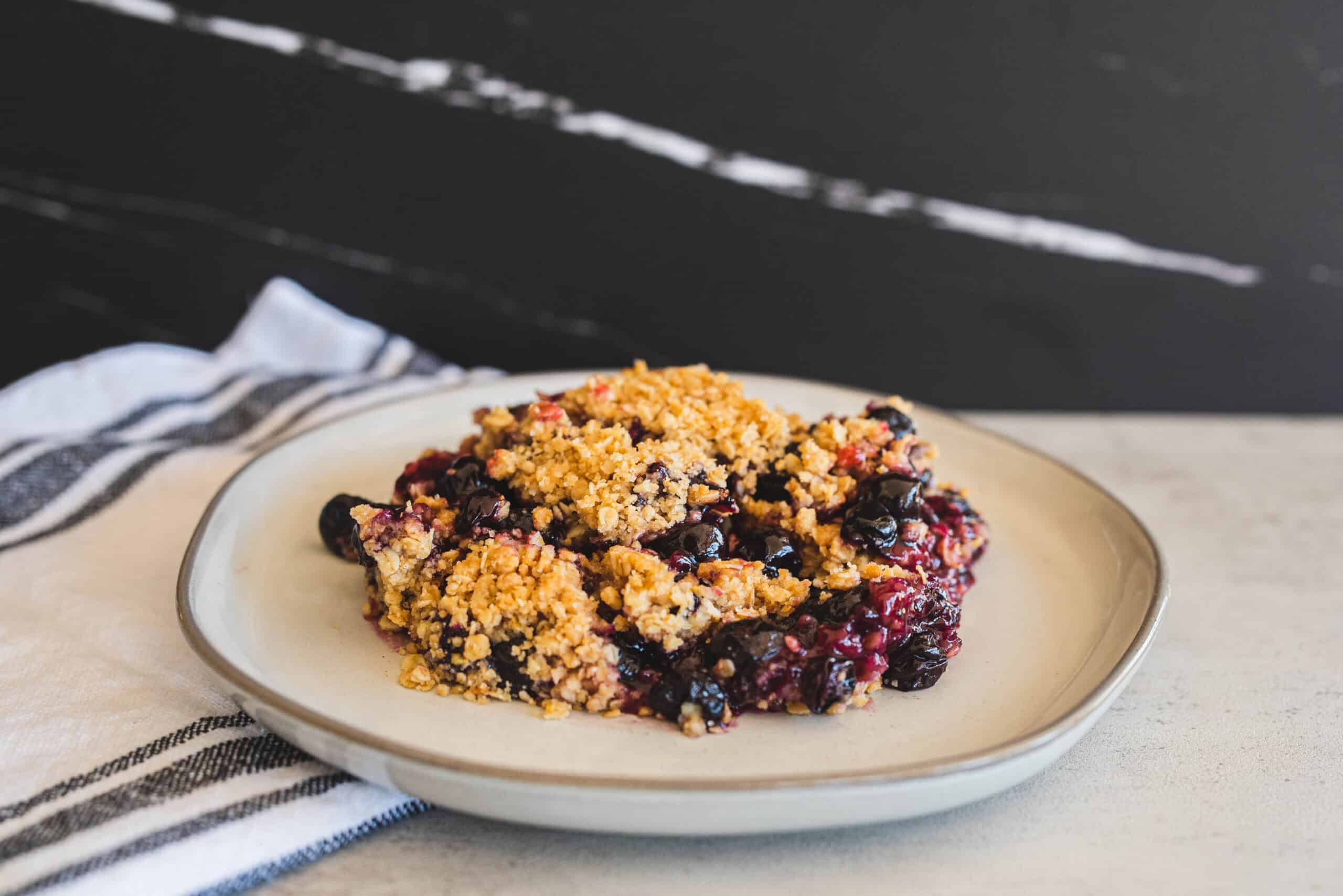 Baked mixed berry crumble on a white plate with a black background and striped cloth, perfect for summer dessert, healthy berry dessert, easy berry crumble recipe.