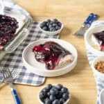Creamy blueberry cheesecake topped with blueberry compote served on white plates with fresh blueberries and chopped nuts, on a rustic wooden table with serving dishes and cutlery.