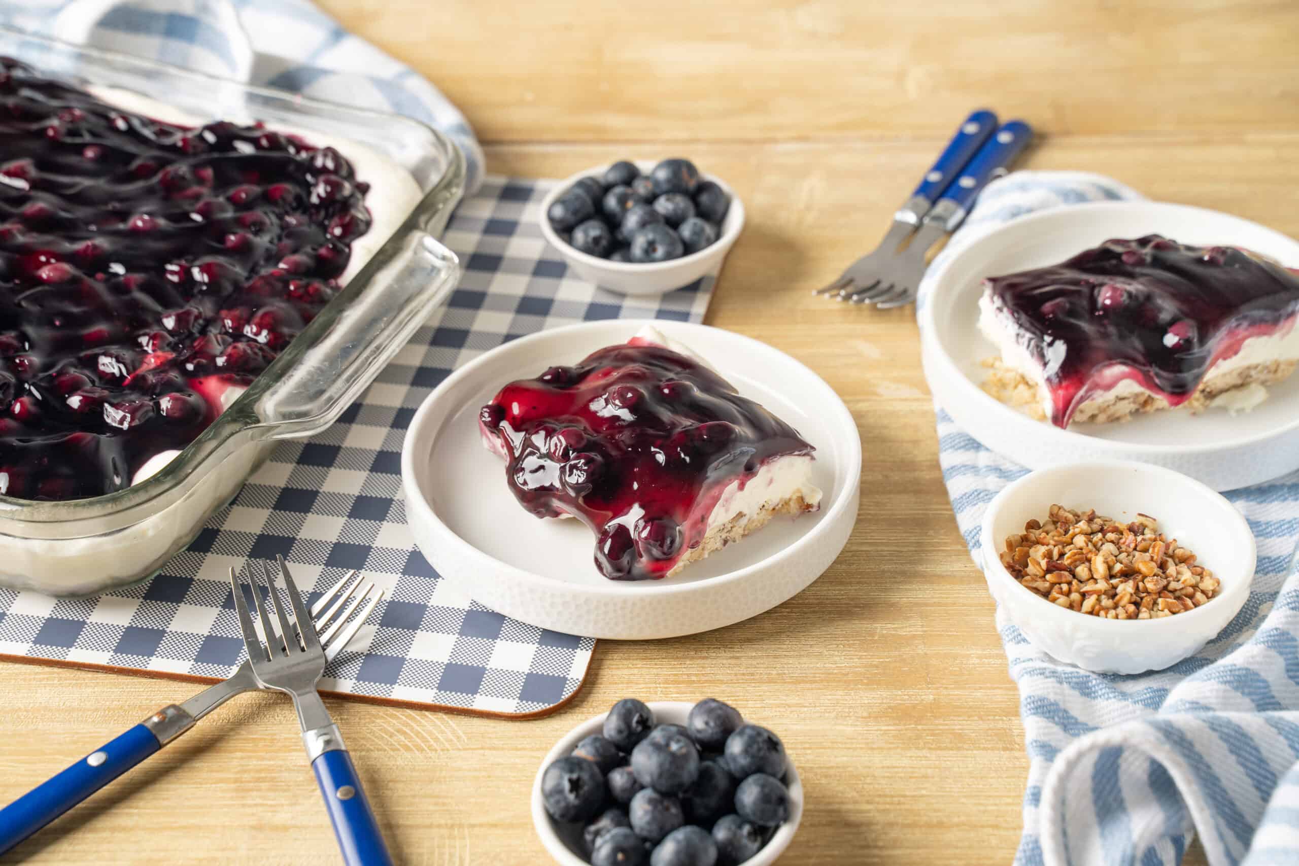 Creamy blueberry cheesecake topped with blueberry compote served on white plates with fresh blueberries and chopped nuts, on a rustic wooden table with serving dishes and cutlery.