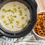 Creamy potato soup topped with chopped green onions in a slow cooker, served alongside a bowl of pretzel bites coated in cheese sauce, on a wooden table.