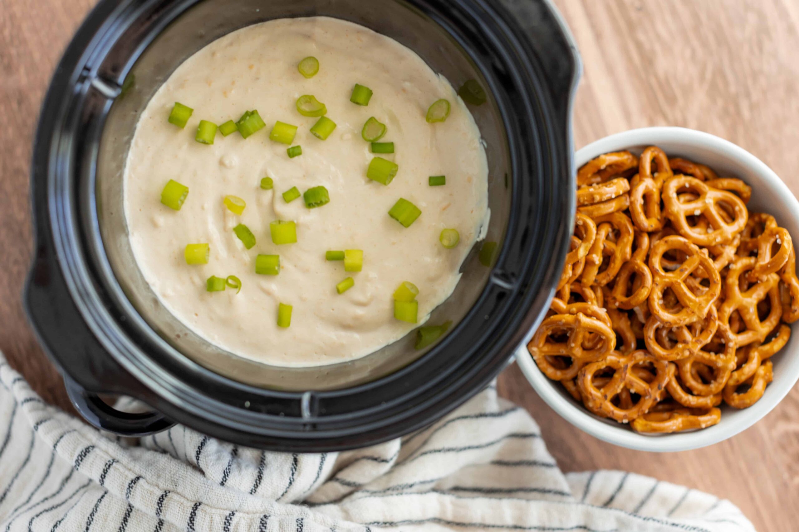 Creamy potato soup topped with chopped green onions in a slow cooker, served alongside a bowl of pretzel bites coated in cheese sauce, on a wooden table.