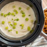 Creamy potato soup topped with chopped green onions in a slow cooker, served alongside a bowl of pretzels for a hearty snack or appetizer.