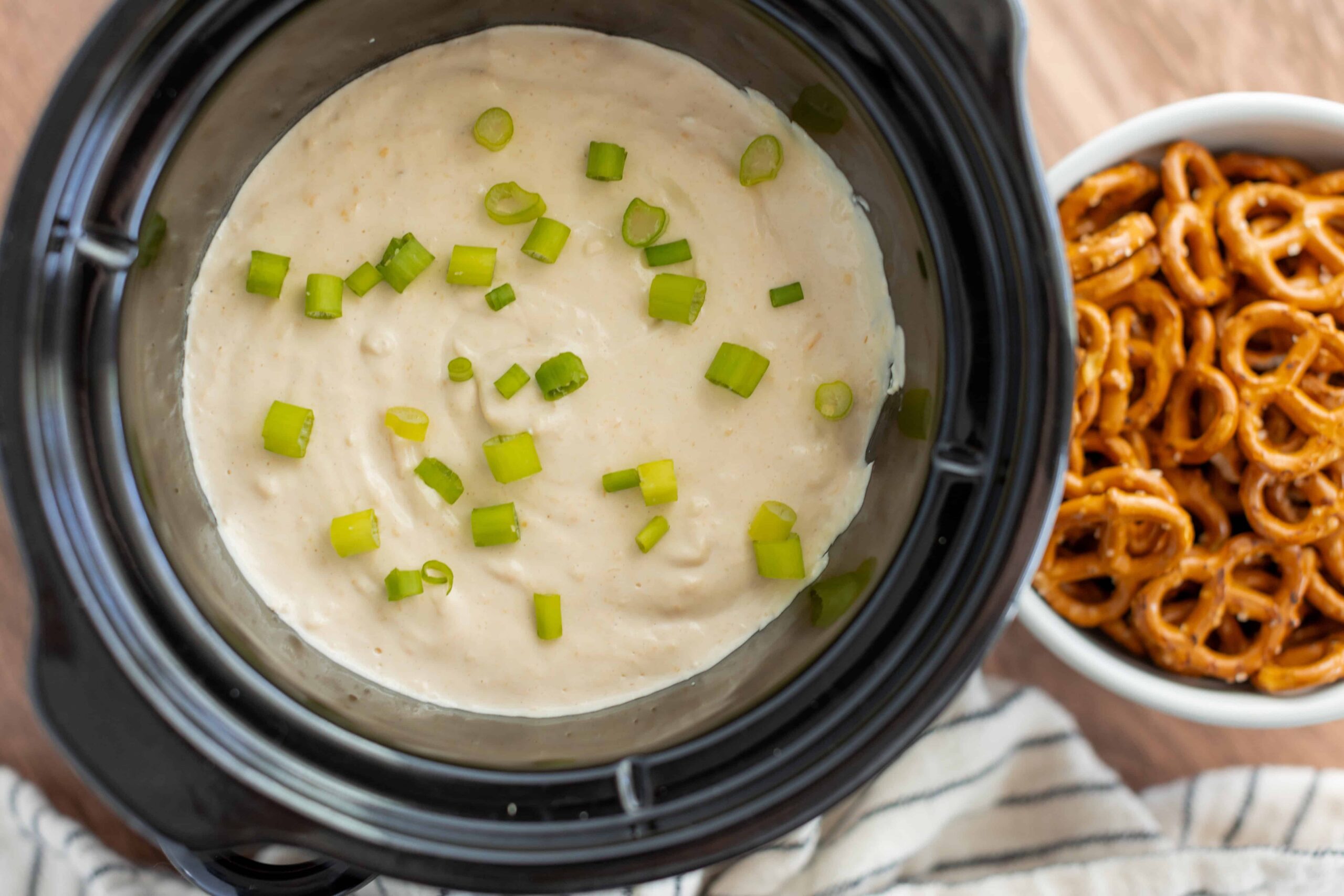 Creamy potato soup topped with chopped green onions in a slow cooker, served alongside a bowl of pretzels for a hearty snack or appetizer.