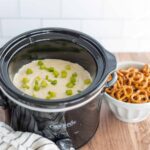 Creamy mashed potatoes with chopped green onions in a black crockpot on a wooden surface, alongside a bowl of freshly cooked, saucy pretzels, perfect for comfort food and snack recipes.