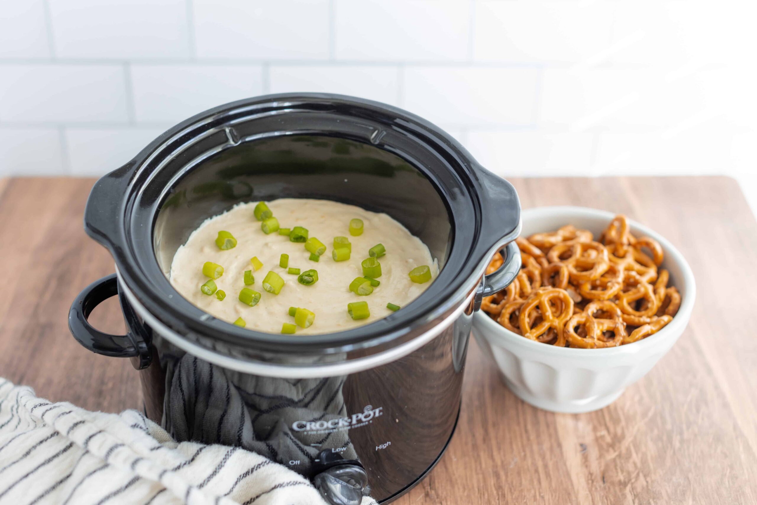Creamy mashed potatoes with chopped green onions in a black crockpot on a wooden surface, alongside a bowl of freshly cooked, saucy pretzels, perfect for comfort food and snack recipes.