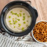 Creamy soup in an Instant Pot garnished with chopped green onions next to a bowl of pretzels on a wooden table, highlighting quick and easy snack recipes.