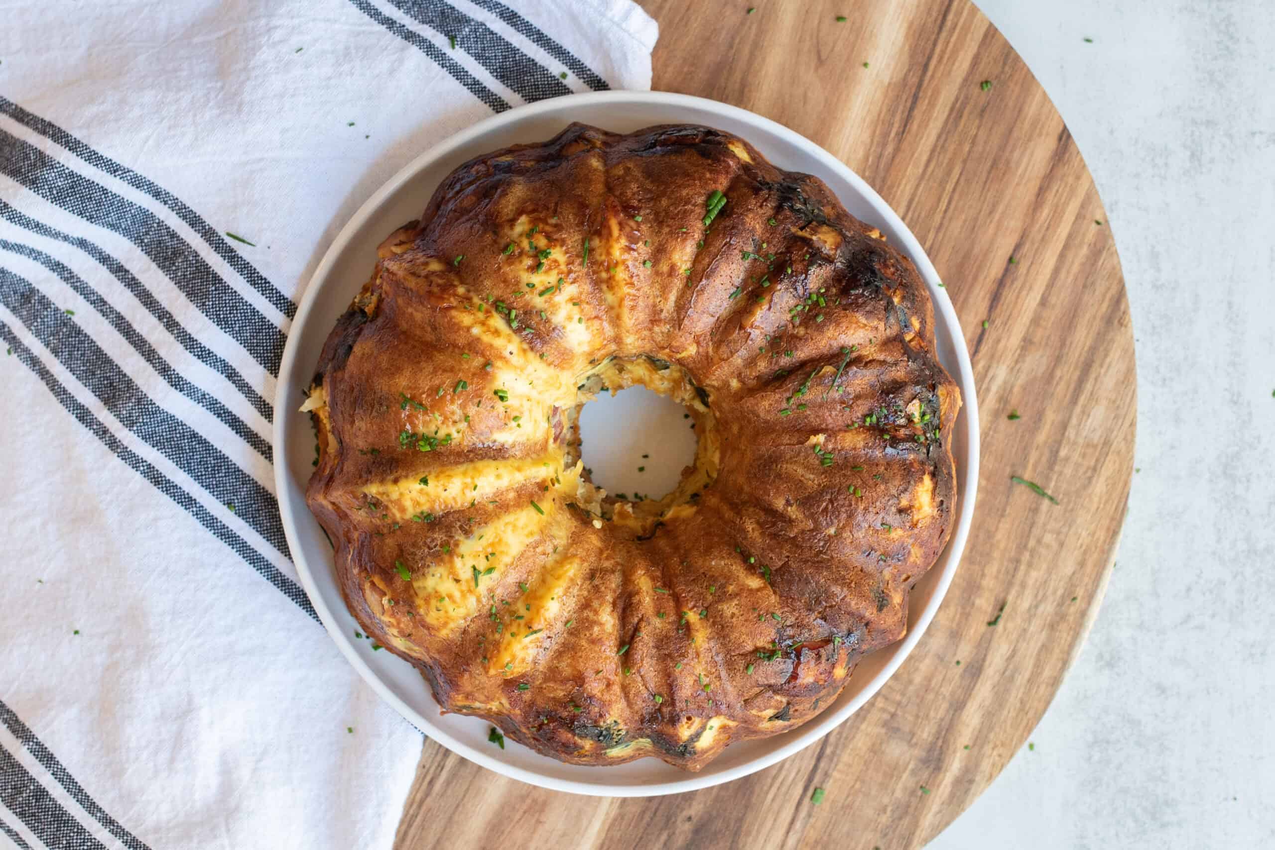 Golden-brown baked Bundt cake garnished with chopped herbs on a white dish, placed on a wooden serving board with a striped cloth, perfect for dessert or tea time.